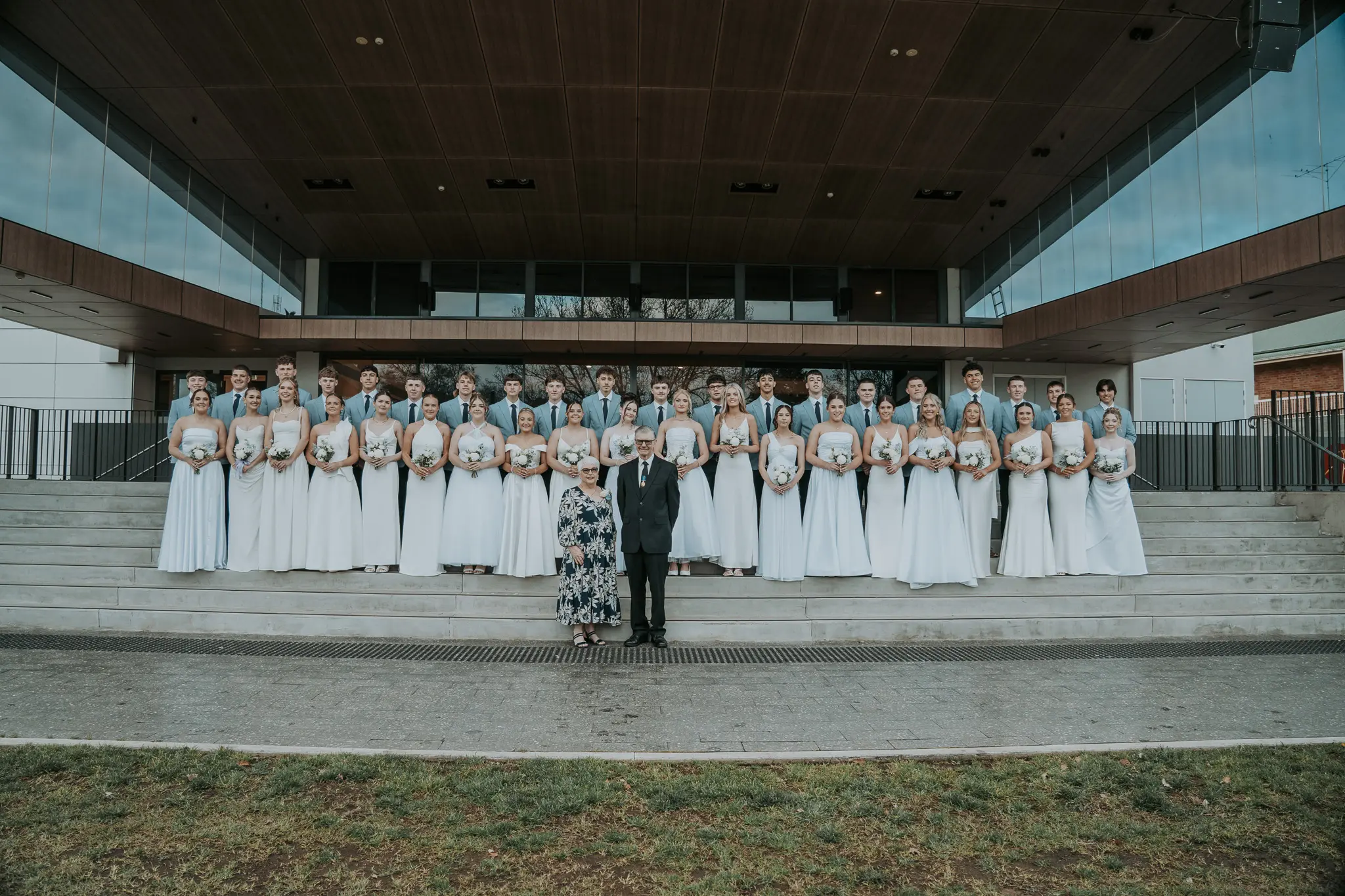 <p>The 20 debutantes and their partners on the steps of the Cooke Park Pavillion with Father Barry Dwyer and matron Pat McCallum for the Parkes Catholic Winter Wonderland Debutante Ball. PHOTOS: Brittney White Photography</p>\\n